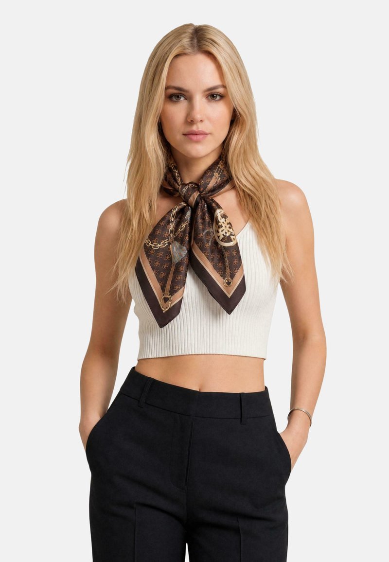 Femme blonde portant un crop top blanc en côte, un pantalon noir et un foulard en soie marron à motifs noué autour du cou, les mains dans les poches.