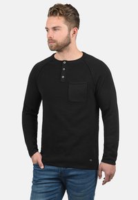 Zwarte lange mouwen henley shirt met geribbelde textuur, drie-knop sluiting en borstzak. Gecombineerd met blauwe jeans.
