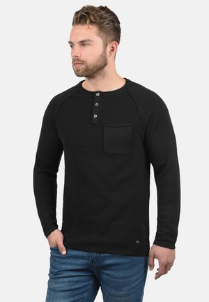 Camisa henley de manga larga negra con textura acanalada, tapeta de tres botones y bolsillo en el pecho. Llevada con jeans azules.
