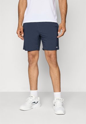 Homme portant un short de sport bleu marine, des chaussures de sport blanches, des chaussettes blanches et un t-shirt blanc, debout devant un fond uni.