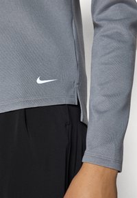 Pilkas sportinis ilgos rankovės marškinėlis su šonine išpjova ir balta Nike logotipu prie krašto, derinamas su juodomis kelnėmis. Lygi, kvėpuojanti medžiagos textura.