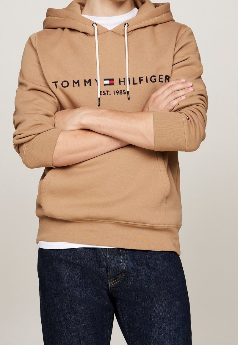 Brun bomullshoodie med en kangoroolficka, justerbara snörningar och broderad logotyp. Matchad med blå denimjeans.