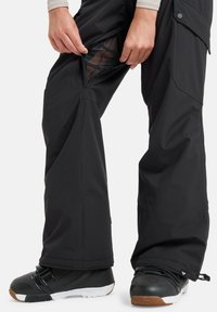 Black ski pants with a zippered mesh ventilation pocket on the thigh. Features a relaxed fit and reinforced hem, paired with black snow boots.