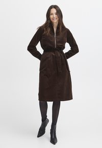Hnedé kordové šaty so zipsom vpredu, dlhými rukávmi a zodpovedajúcim opaskom. Ankle boots dotvárajú vzhľad. Hladká textúra a dĺžka po stred nôh.