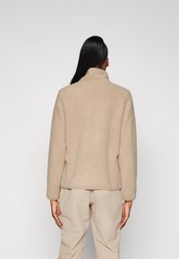 Giacca in pile beige con colletto alto e maniche lunghe, caratterizzata da un tessuto morbido e testurizzato. Indossata con pantaloni beige chiaro, vista da dietro.