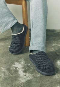 Black and gray checked slippers with a soft interior, paired with gray sweatpants and dark green socks, resting on a textured gray surface.