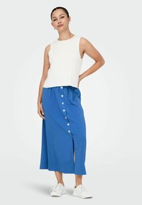 Top bianco a costine abbinato a una gonna midi blu con chiusura a bottoni frontale e spacco laterale, completato da sneakers bianche.