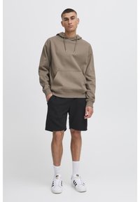 Sweat à capuche marron avec poche avant, cordon de serrage et poignets côtelés, associé à un short noir. Des chaussettes blanches et des baskets à motifs complètent le look.