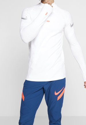 Weißes langärmliges Performance-Shirt mit strukturierten Linien und orangefarbenen Akzenten, kombiniert mit blauen Hosen, die ein großes Logo zeigen.