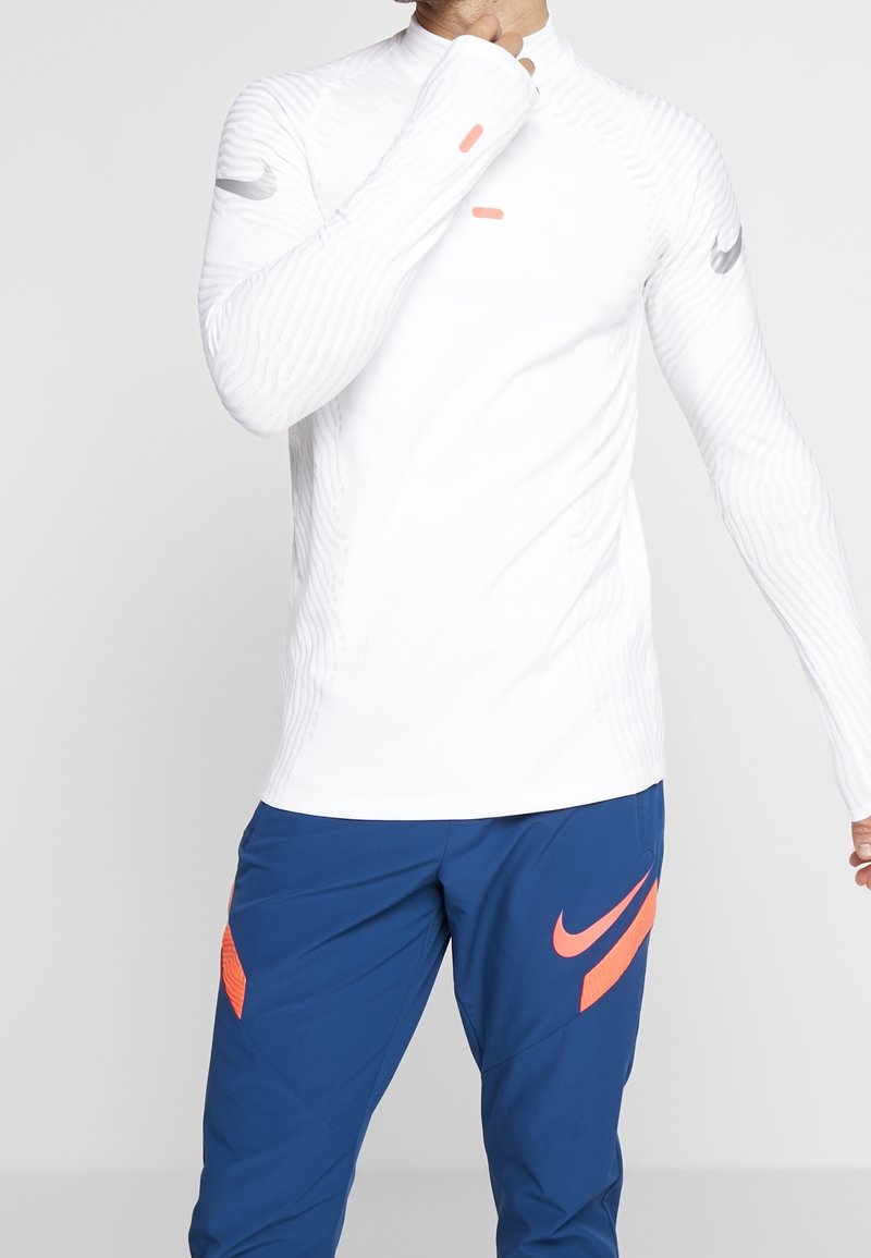 Biała, długorękawowa koszulka sportowa z teksturowanymi liniami i pomarańczowymi akcentami, zestawiona z niebieskimi spodniami z dużym logo.