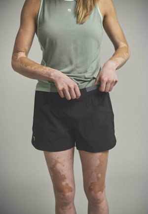 Person mit Vitiligo, die schwarze Sportshorts anpasst, trägt ein ärmelloses hellgrünes Oberteil, gezeigt vom Hals bis zur Mitte des Oberschenkels vor grauem Hintergrund.