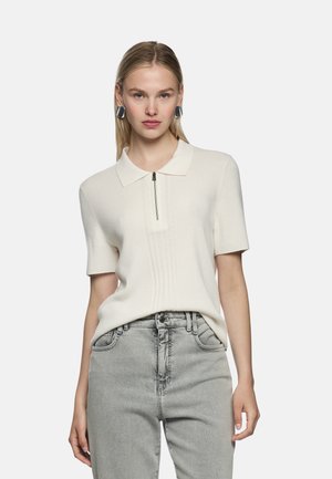 Weißer kurzärmliger Polopullover mit Reißverschluss am Ausschnitt und gerippter Textur, kombiniert mit hellgrauen Jeans. Einfaches, modernes Design.