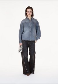 Camicia in denim di colore azzurro chiaro, modello con bottoni, abbinata a jeans a gamba larga scuri con cuciture bianche. La modella tiene una borsa metallica.