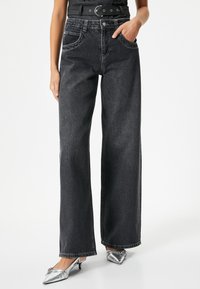 Jeans negros de tiro alto y pernera ancha con bolsillos delanteros, costuras en contraste y una suave textura de denim, combinados con tacones plateados de punta afilada.