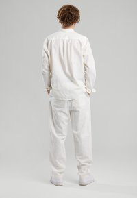 Una persona con capelli ricci sta in piedi di spalle, indossando una camicia bianca ampia, pantaloni bianchi e sneakers bianche su uno sfondo grigio semplice.