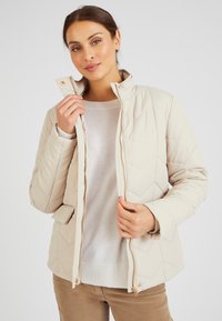 Giacca trapuntata beige con colletto alto, chiusura con zip e dettagli dorati. Presenta un motivo a rombi testurizzato e tasche laterali.