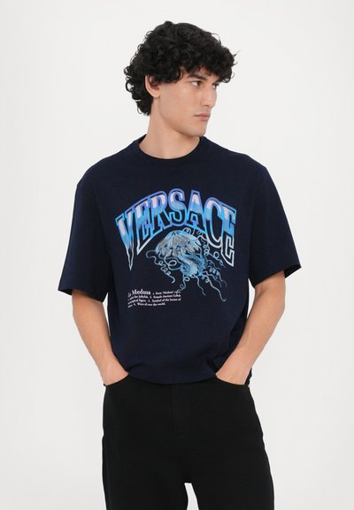Tumesin lühikeste varrukatega T-särk puuvillast, millel on julge graafiline trükk, kus on kirjas "VERSACE" ja Medusa kujundus sinises, koos alloleva tekstiga.