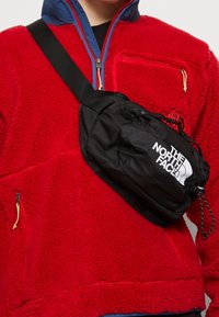 Bolsa de nylon negra tipo bandolera con logo, llevada sobre una chaqueta de forro polar roja. Cuenta con correa ajustable y cierres de cremallera.