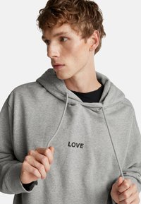 Giovane con capelli ricci che indossa un felpa grigia con la scritta "LOVE", tiene i lacci della felpa e guarda a sinistra su uno sfondo neutro.