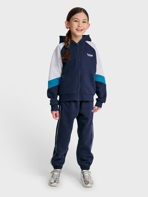 Junges Mädchen mit geflochtenem Haar, das einen marineblauen Hoodie mit grauen und türkisfarbenen Ärmeln, passende marineblaue Jogginghosen und silberne Sneakers trägt, lächelnd und stehend.