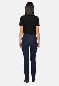 Jeans ajustados de mezclilla azul oscuro con una textura suave, cinco bolsillos, una cintura alta y un parche de cuero marrón en la parte trasera. Camiseta negra puesta.
