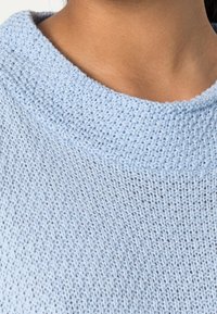 Pull tricoté bleu clair avec un motif texturé, présentant un large col arrondi et un tissu doux. Vue en gros plan du col et de l'épaule.
