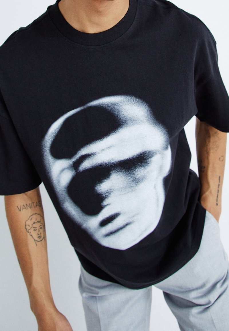 Black cotton t-shirt featuring a large white graphic of a face. Crew neck and loose fit. Visible tattoos on the wearer’s arm.