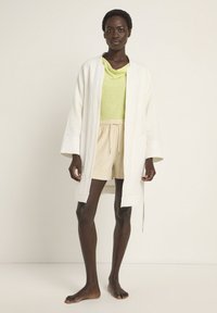 White textured kimono-style jacket, layered over a lime green top and beige shorts with drawstring, styled with bare feet.