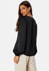Blusa nera in satin con maniche lunghe a palloncino e polsini elastici, con una texture liscia e una vestibilità rilassata, abbinata a pantaloni in colori neutri.
