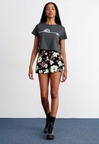T-shirt grigio accorciato con un design semplice abbinato a pantaloni shorts neri con stampa floreale. Il modello indossa stivali neri con lacci. Sfondo bianco uniforme.