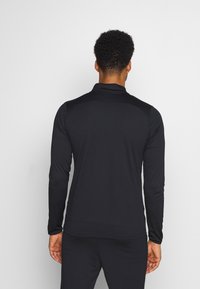 Zwarte sportieve langslavend shirt met een kraag, gladde textuur, slim fit ontwerp en stiksels langs de schoudernaden. Achteraanzicht weergegeven.