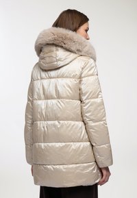 Giacca imbottita beige dal tessuto lucido, con un ampio cappuccio foderato in pelliccia e cuciture orizzontali sul corpo per un maggiore calore.