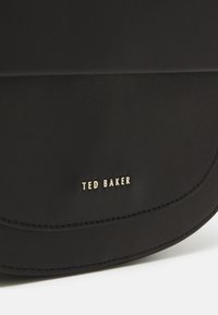 Musta nahkalaukku, jossa on kaareva muotoilu ja kultaiset "TED BAKER" -brändäykset. Sileä pinta, jossa on näkyvät sauman yksityiskohdat.