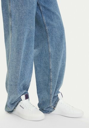 Persona con pantalones vaqueros azules holgados y zapatillas blancas Reebok, de pie sobre un suelo de color claro.