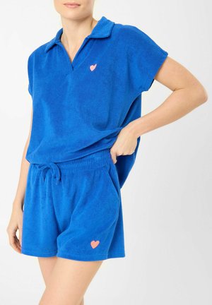 Femme portant une chemise bleue à col en tissu éponge assorti et un short à cordon avec de petits détails de cœurs roses sur le haut et le short.
