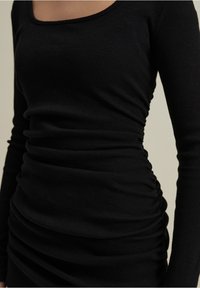 Černé žebrované šaty s dlouhým rukávem, kulatým výstřihem a nabíranými detaily po stranách, které zvýrazňují strukturu a přiléhavý design.