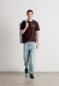 Camiseta de algodón marrón con texto impreso, jeans de mezclilla azul claro y zapatos negros. Una chaqueta azul claro está colgada sobre un hombro.