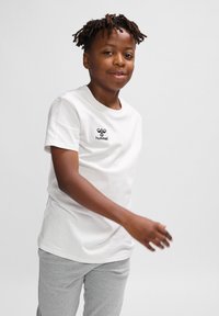 Hvid T-shirt med korte ærmer, der har et sort "hummel" logo på venstre bryst, parret med grå sweatpants. Glat bomuldsstruktur.