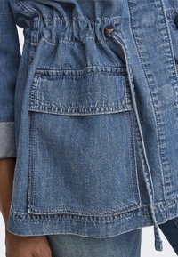 Veste en denim bleu clair avec taille ajustable à cordon, deux poches avant et texture douce. Présente des détails de couture visibles.