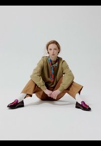 Jeune personne aux cheveux rouges assise par terre, jambes écartées, portant un pull beige, un pantalon marron, des chaussettes blanches, une écharpe colorée et des chaussures bordeaux brillantes.