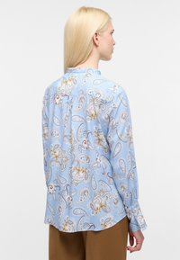 Camicia in poliestere azzurro chiaro con motivo paisley bianco, oro e marrone. Presenta maniche lunghe, una piega sul retro e polsini con bottoni.