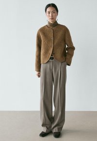 Brauner, flauschiger Cardigan mit rundem Ausschnitt und fünf Knöpfen, getragen über einem braunen Rollkragenpullover. Kombiniert mit hellbraunen, weiten Hosen.