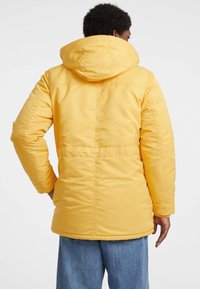 Geel geïsoleerde jas met capuchon, met een rechte snit, getailleerde textuur en zichtbare stiknaden op de achterkant.