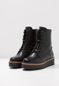 Botas de cuero negro con cordones, con una gruesa suela de plataforma negra y un acento de goma natural texturizada. Presentan un acabado suave y una puntera redondeada.