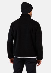 Czarna polarowa kurtka z wysokim kołnierzem i gładką warstwą materiału na rękawach. Noszona z czapką z dzianiny w czarnym kolorze, która ma fakturowaną powierzchnię.