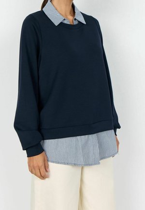 Kvinde iført marineblå sweatshirt over en blå- og hvidstribet skjorte med krave og beige bukser, stående mod en ensartet lys baggrund.