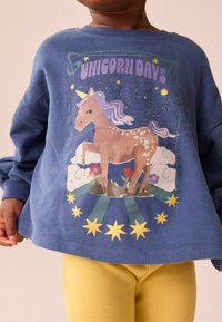 Niebieska bluza z kapturem z ilustracją jednorożca i napisem "DNI JEDNOROŻCA". Złote gwiazdy i kwiatowe akcenty zdobią wzór. Wyświetlono żółte legginsy.