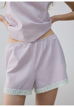 Femeie purtând pantaloni scurți de bumbac mov deschis cu dantelă albă și un top lejer asortat, cu mâinile sprijinite pe șolduri.