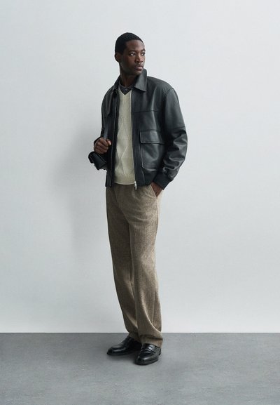 Giubbotto bomber in pelle nera, maglione lavorato a maglia color crema, pantaloni marroni testurizzati e scarpe nere, in piedi contro uno sfondo grigio chiaro.