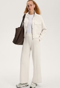 Weiße gekürzte Jacke mit zwei Taschen, weißes enganliegendes Oberteil, weit geschnittene weiße Hose und graue Sneakers; braune Tote-Bag mit Schultergurt.